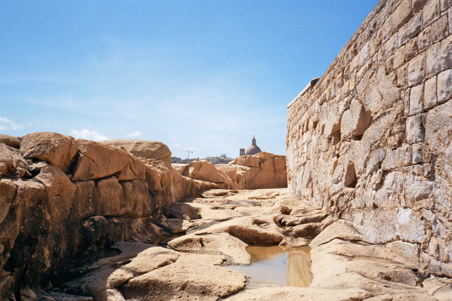 Sonniger Pfad zwischen rauen Felsen und einer historischen Kalksteinmauer auf Malta mit einer Kirchenkuppel im Hintergrund.