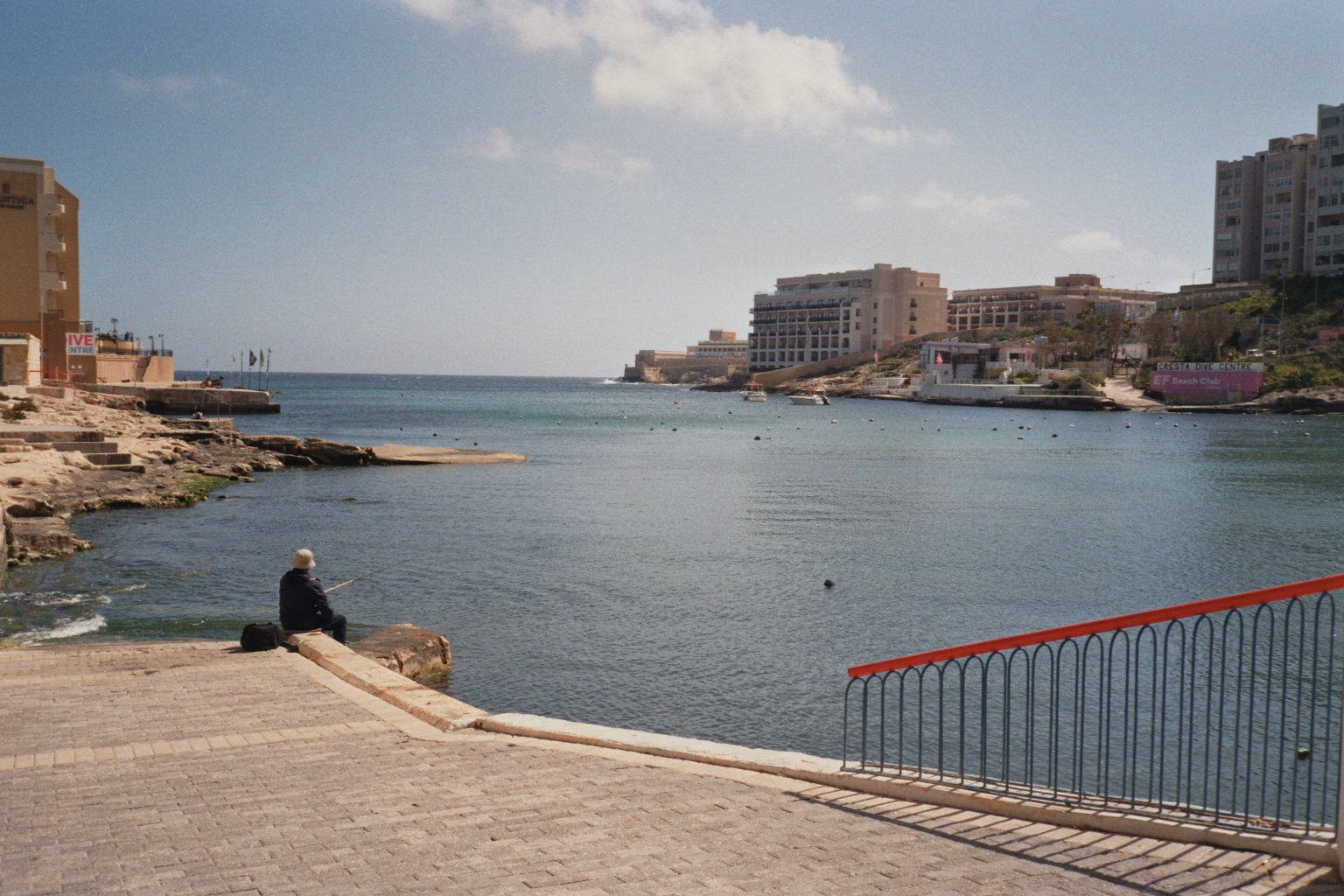 Angler an einer Uferpromenade auf Malta mit Blick auf eine Meeresbucht und Hotelgebäude.